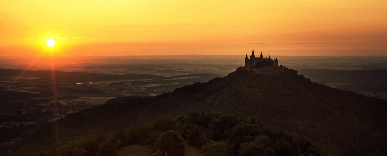 Burg Hohenzollern thront majestätisch auf einem Hügel. Die Sonnenstrahlen beleuchten die Landschaft, während der Himmel in sanften Orange- und Gelbtönen erstrahlt. Umgeben von Wäldern, wirkt die Burg gleichzeitig imposant und romantisch.