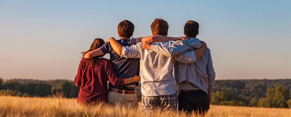 Vier junge Menschen stehen mit dem Rücken zu uns in einem Gerstenfeld, haben die Arme übereinander gelegt und schauen auf den Horizont. Es ist ein sonniger Tag, im Hintergrund ist ein Wald zu sehen.
