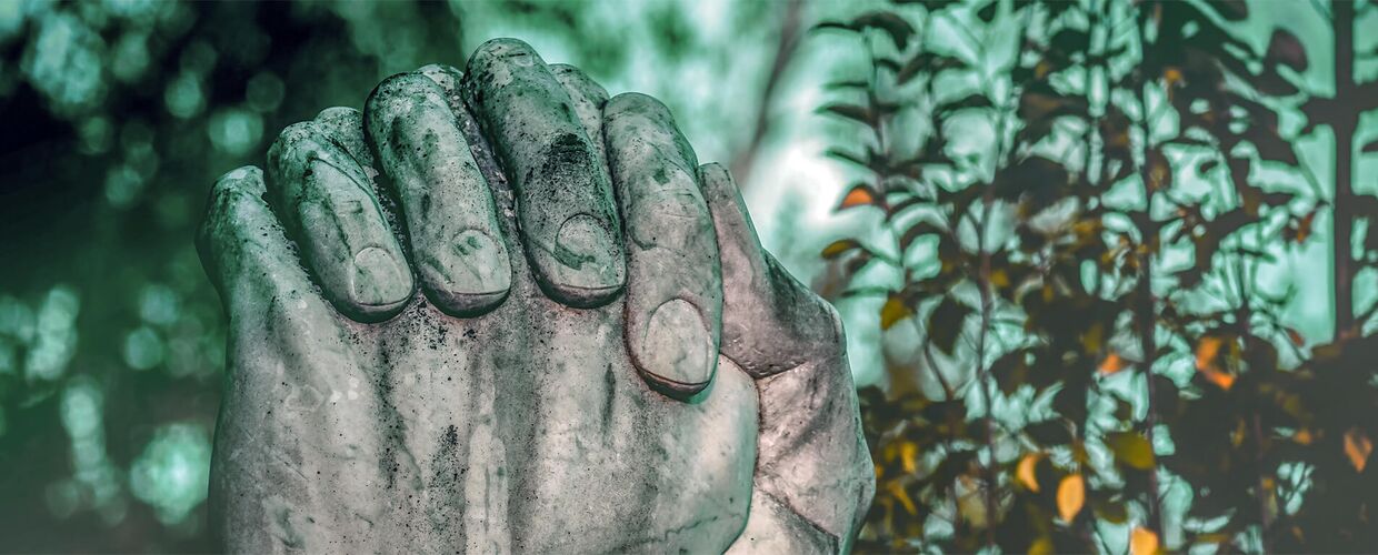 Zwei große, steinerne Hände sind betend gefaltet. Der graue Stein ist verwittert und wirkt alt. Im Hintergrund sieht man unscharfe Bäume mit grünem und herbstlich gefärbtem Laub. Licht fällt hindurch und schafft eine ruhige, spirituelle Stimmung.