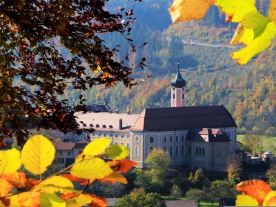 Das Kloster Beuron liegt malerisch in einer sanften Hügellandschaft, umgeben von buntem Herbstlaub. Die Sonne beleuchtet die imposante Klosteranlage mit ihrem markanten Kirchturm, Wälder und Hügel verstärken die ruhige Atmosphäre des Ortes.