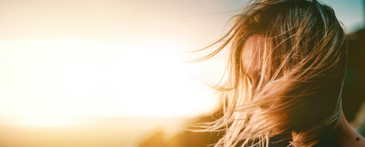 Kopf einer jungen Frau, deren Haare vom Wind verweht werden. Man sieht sie von der Seite, sie blickt nach unten. Im Hintergrund scheint die Sonne.